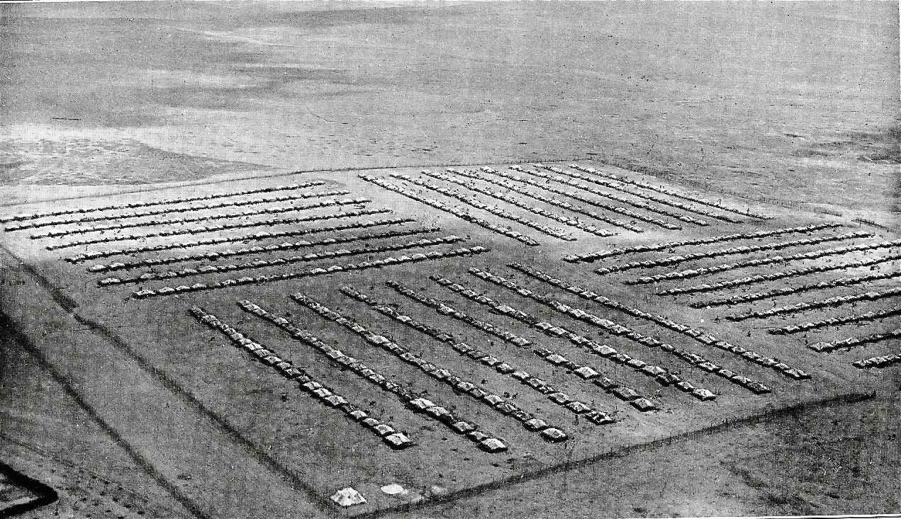 A black and white photograph taken from above of a concentration camp. It is a series of small huts arranged in lines surrounded by a fence and watch towers.